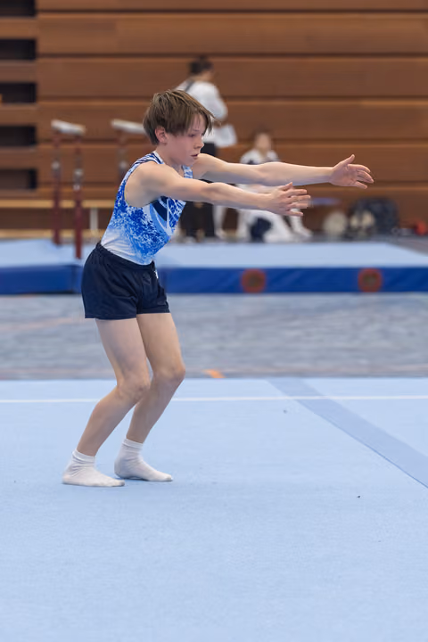 Young gymnast in blue leotard performs expressive arm movements during floor routine, displaying intense focus and concentration