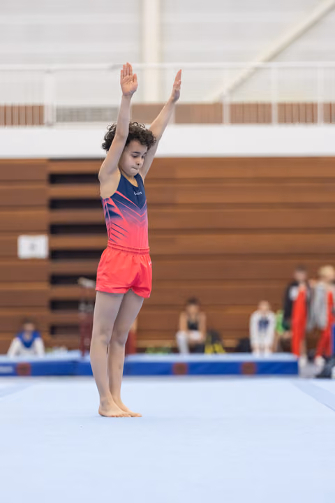 Young gymnast stands with arms raised overhead on floor mat, focused expression, preparing for routine in sports hall