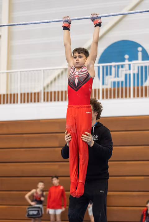 Young gymnast in red outfit hangs from horizontal bar with focused expression while coach spots from below