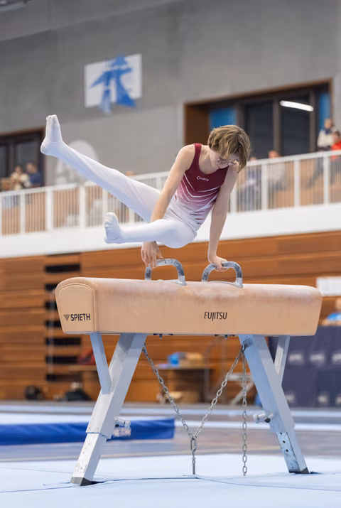 Gymnast performs impressive straddle hold on pommel horse, legs extended horizontally while balancing on handles in training facility