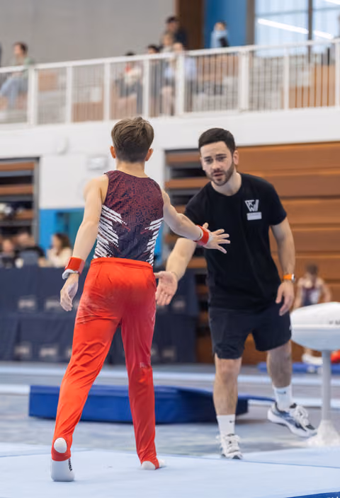 Coach in black gestures toward a gymnast wearing a sparkly leotard and red pants on the competition floor
