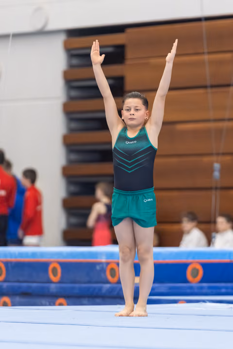Young gymnast in teal and black leotard stands with arms raised overhead on floor exercise mat, showing focused expression