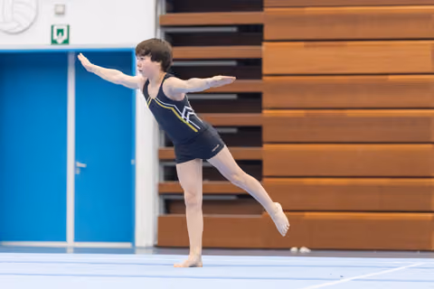 Young gymnast performing an arabesque during floor routine, arms extended gracefully in navy leotard at indoor training facility