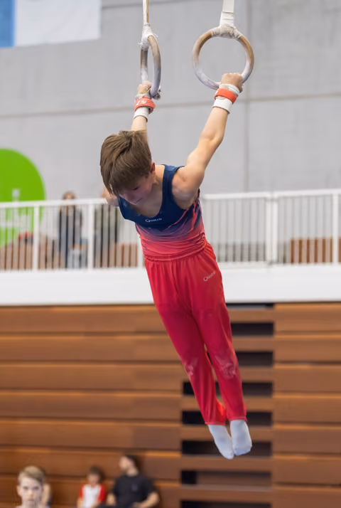 Young gymnast in pink pants and blue leotard hanging from gymnastics rings during practice in an indoor gym facility
