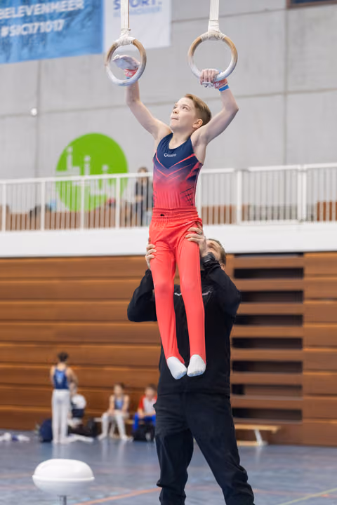 Young gymnast in red gradient leotard performs ring routine while coach spots from below in indoor training facility