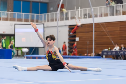 Young gymnast performs a split on the floor with one arm gracefully extended, displaying flexibility and poise in the arena