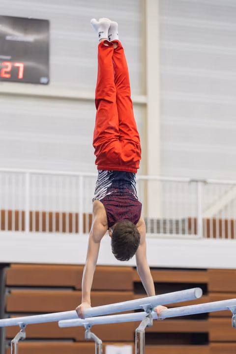 Young gymnast performs a handstand on parallel bars during training, wearing red hoodie and purple shorts in indoor gym