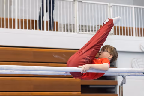 Young gymnast in red performs on horizontal bar, legs extended upward, in indoor gymnasium