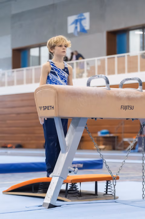 Young gymnast with blonde hair stands beside pommel horse, gazing upward with focused expression in training facility
