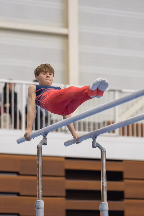 Young gymnast performing horizontal hold on parallel bars, legs extended in red pants, focused expression in training facility