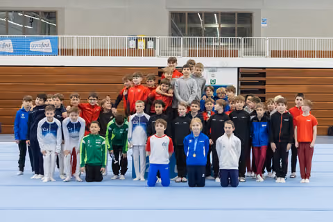 Large group of young gymnasts in colorful tracksuits posing together on blue mat in gymnasium with wooden bleachers