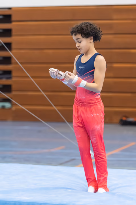Young gymnast in pink and navy leotard carefully adjusts white wrist guards while standing on blue mat in gymnasium