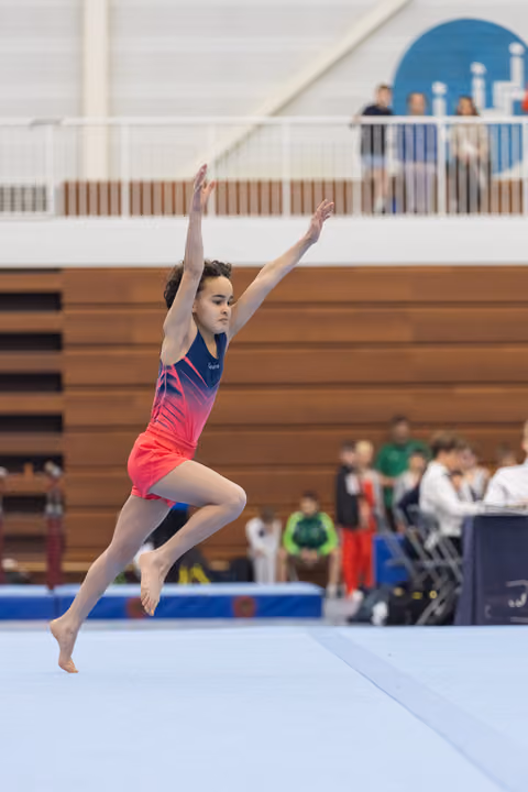 Young gymnast performs graceful floor routine with arms extended upward, wearing vibrant pink-to-blue gradient leotard