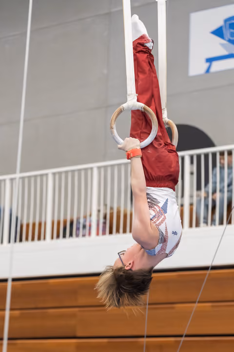 Young gymnast hangs upside down on still rings, demonstrating strength and control in a gym with spectators in background