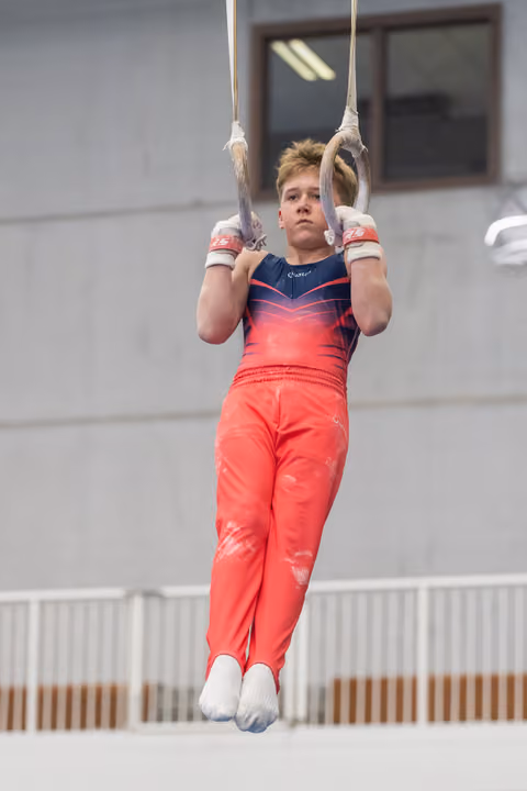 Young gymnast in coral outfit performing still rings routine, displaying intense focus and strength during apparatus work