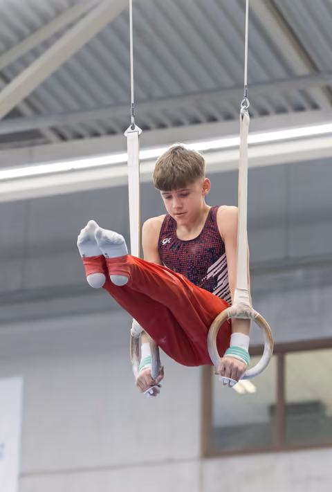 Young gymnast performing horizontal hold on rings, wearing purple leotard and red pants in indoor training facility