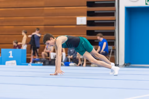 Young gymnast in teal and green uniform performs a deep lunge stretch on floor mat during warmup, focused and preparing