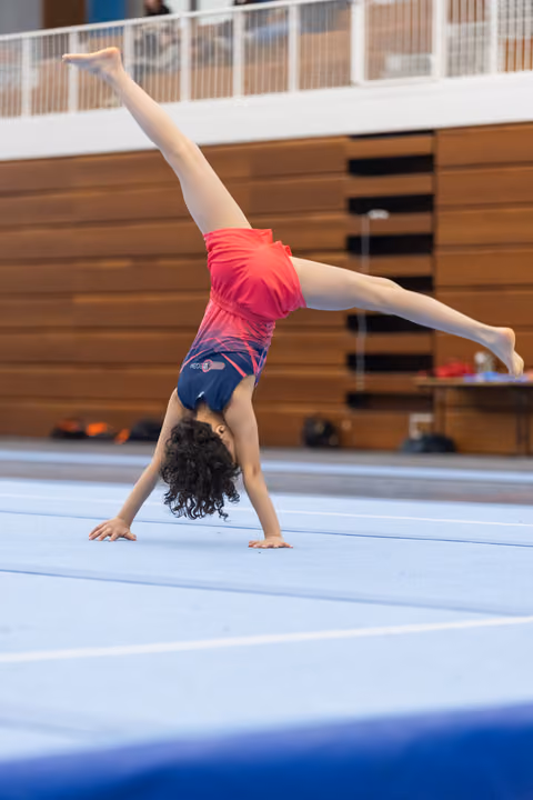 Young gymnast performing handstand on blue floor mat, pink leotard, curly hair, training in gymnasium