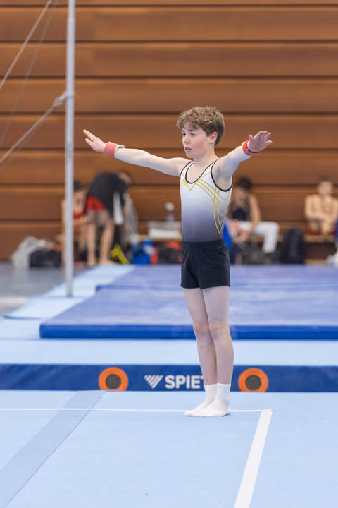 Young male gymnast performing floor routine with arms extended wide, showing focus and determination in gym with wooden walls