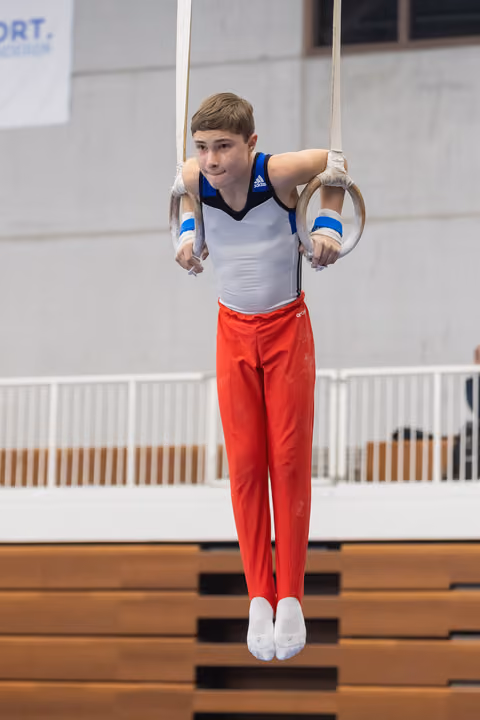 Young male gymnast performing on still rings in white and red uniform, displaying focused concentration during apparatus work