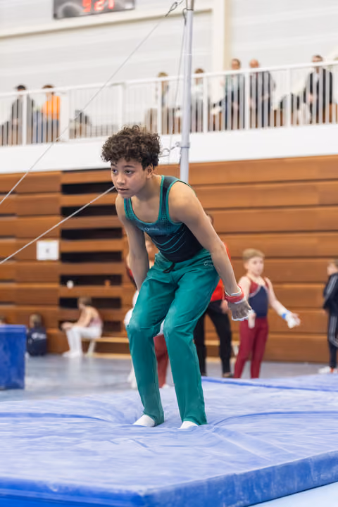 Young gymnast in teal uniform performs floor routine with focused expression in gymnasium with spectators in bleachers
