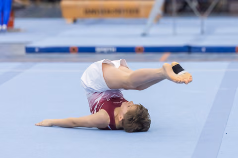 Young gymnast performs floor routine with back on mat and legs extended upward, demonstrating flexibility and control