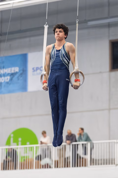 Male gymnast suspended on rings in focused concentration, wearing navy uniform with white hand guards during routine