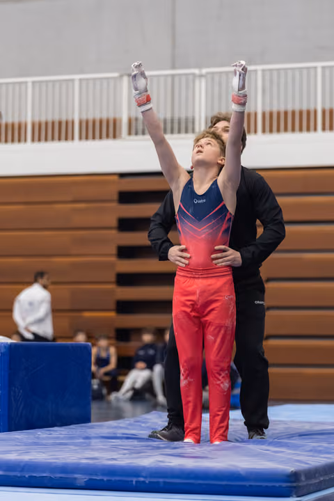Young gymnast in red ombre outfit raises arms in triumph while coach stands supportively behind on blue mat in gymnasium