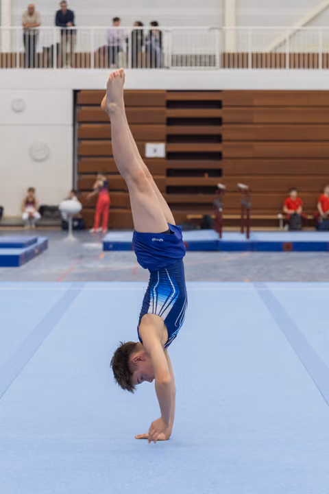 Young gymnast performs a handstand on floor mat, legs extended upward, with spectators watching from bleachers