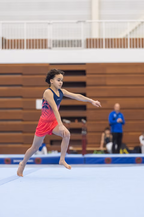 Young gymnast performs floor routine with extended arms, displaying focused concentration in gymnasium with bleachers