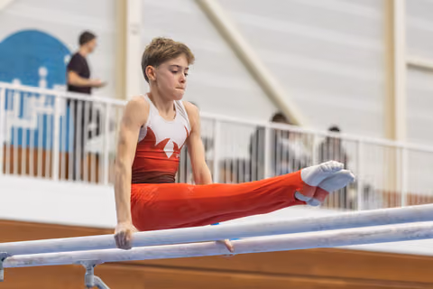 Young gymnast in red and white uniform performing L-sit position on parallel bars with concentrated expression
