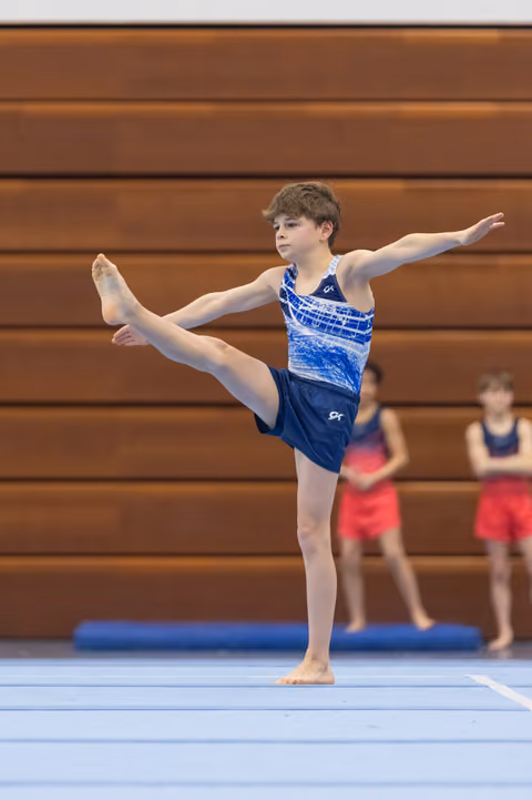 Young gymnast executes a standing vertical leg hold with extended arms during floor routine at indoor gymnastics meet