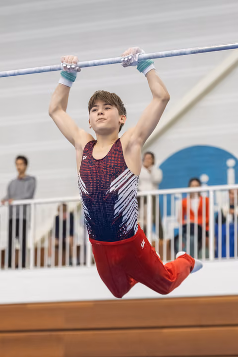 Young gymnast hanging from high bar with focused expression, wearing red and blue uniform with hand grips at indoor training facility