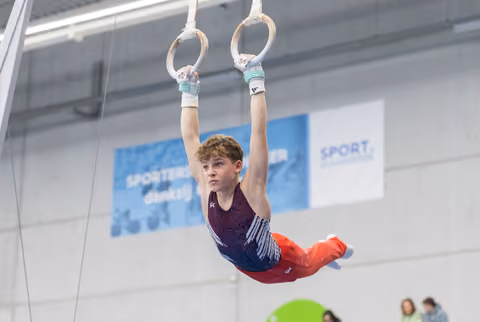 Young gymnast performs on still rings, body horizontal mid-routine, focused expression in bright training facility