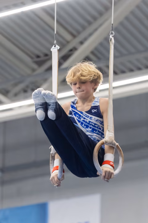Young gymnast with blonde hair performs L-sit on rings, displaying focus and strength during training in indoor facility