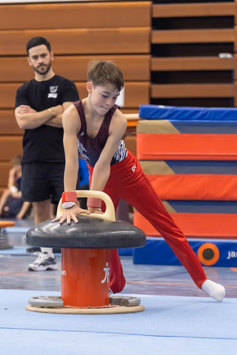 Young gymnast in burgundy leotard and red pants practices on pommel horse while coach observes in training facility