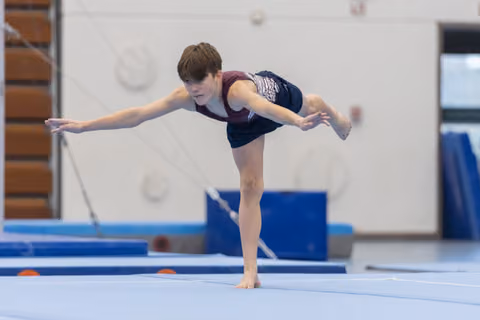 Young gymnast performs arabesque balance on floor exercise mat, arms extended with focused concentration in training facility