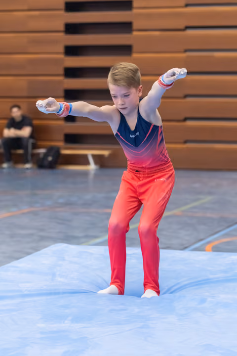 Young male gymnast in gradient pink uniform performs floor routine with arms extended, showing focused concentration
