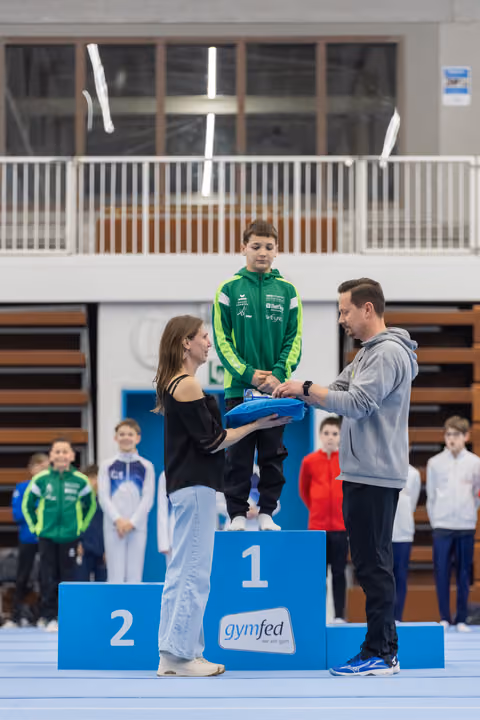Boy in green tracksuit stands on first place podium while officials present medal at gymnastics awards ceremony