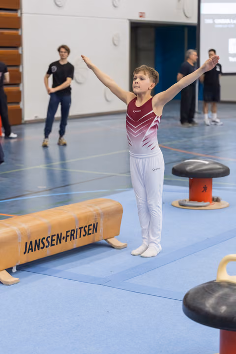 Young gymnast strikes triumphant pose with arms raised after routine, while teammates observe in background