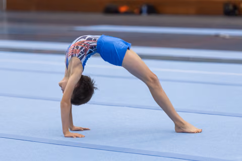 Young gymnast performs a backbend on floor mat, displaying flexibility and strength in blue patterned leotard