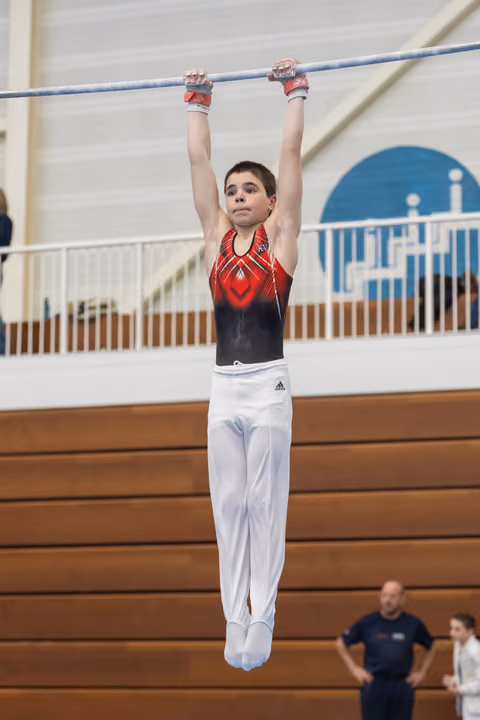 Young gymnast in red and black leotard hangs from high bar with focused expression during training session