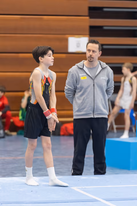 Young gymnast in black uniform stands attentively beside coach in grey jacket on blue floor mat during training session