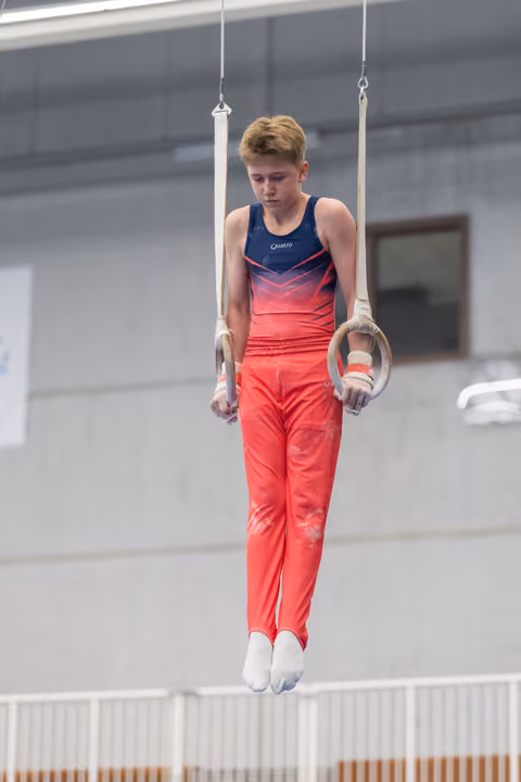 Young male gymnast performs on rings, displaying intense concentration while suspended in the air in gradient outfit
