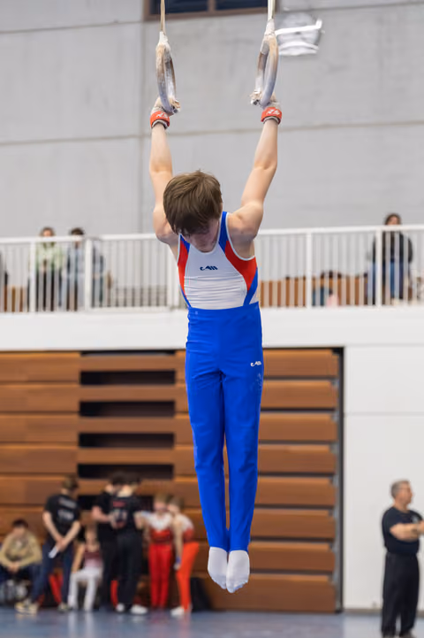 Young gymnast in blue and red uniform performs vertical hold on rings, demonstrating strength and focus in gymnasium