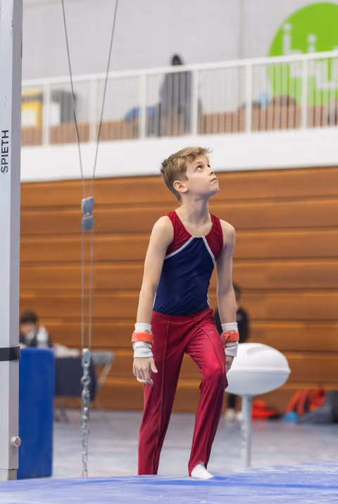 Young gymnast in red and navy leotard gazes upward after completing high bar routine, showing focused concentration
