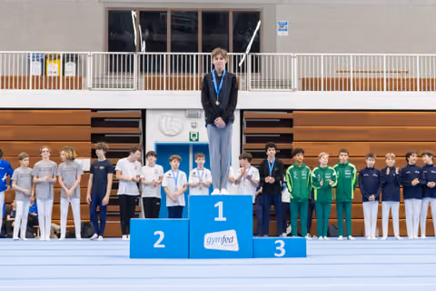 Young gymnast standing on first place podium wearing medal, with second and third place teams lined up beside podium
