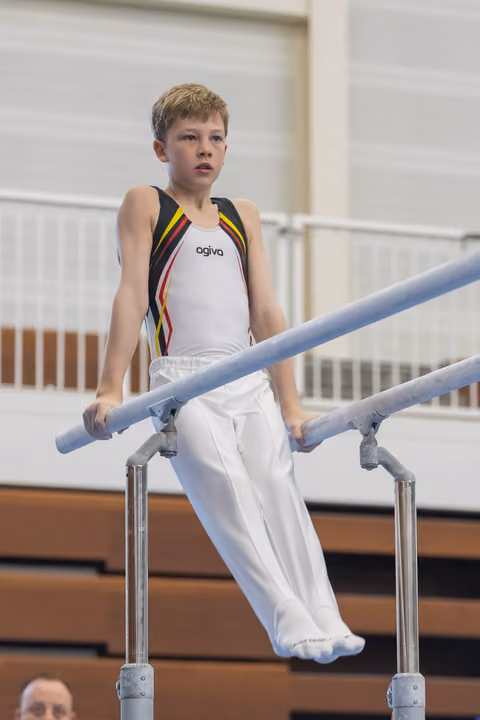 Young gymnast in white and rainbow-striped leotard performs on parallel bars with concentrated expression in gymnasium