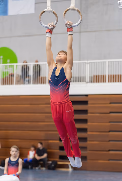 Young gymnast in gradient pink outfit hangs from rings with focused expression during training session in indoor gymnasium