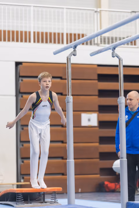 Young male gymnast lands on orange springboard with arms outstretched, coach in blue observing nearby in gymnasium
